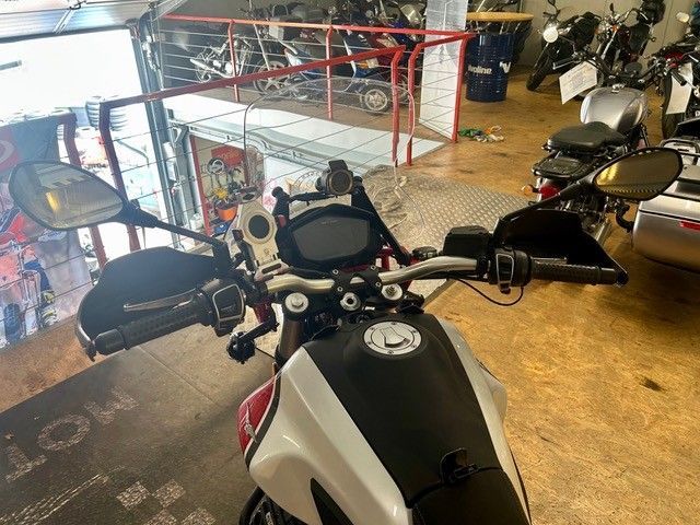 Ein Motorrad ist in einem Ausstellungsraum oder einer Garage geparkt; im Hintergrund sind weitere Motorräder und ein rotes Geländer zu sehen.