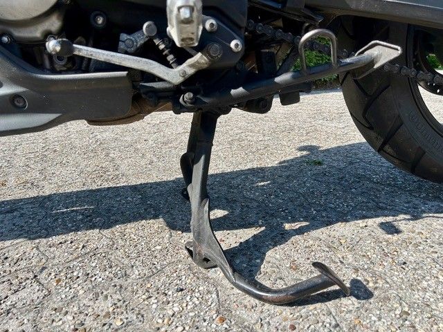 Ein verbogener Motorradständer liegt auf einer asphaltierten Straße neben dem Hinterrad.