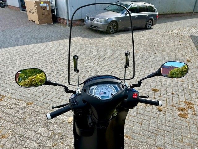 Ein Motorroller mit Windschutzscheibe ist auf einer gepflasterten Auffahrt neben einem Auto geparkt.