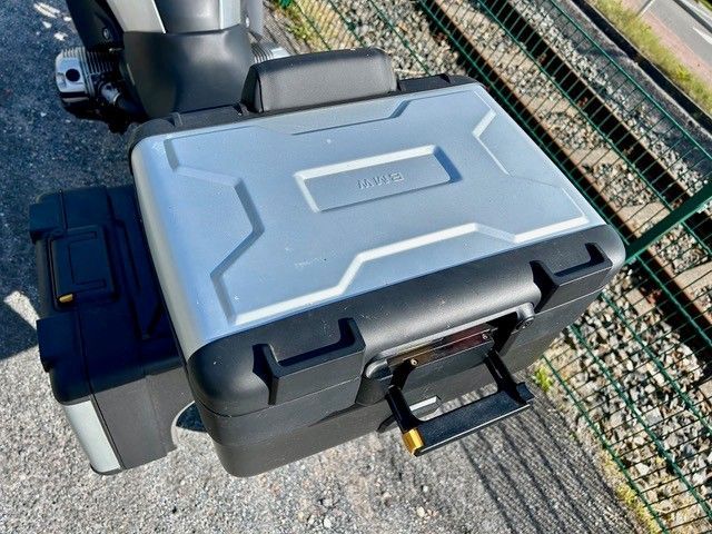 Silbernes und schwarzes Motorrad-Topcase, Griff nach unten, vor einem grauen Hintergrund in der Nähe eines grünen Zauns.