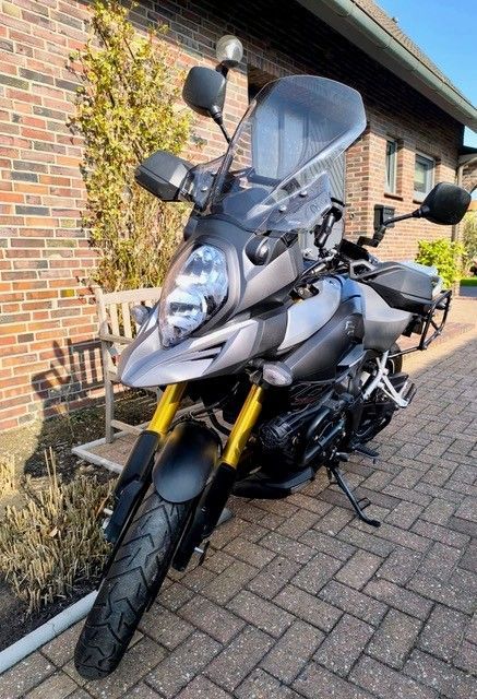 Silbernes und schwarzes Suzuki V-Strom Adventure-Motorrad, geparkt auf einer gemauerten Auffahrt vor einem Backsteingebäude.