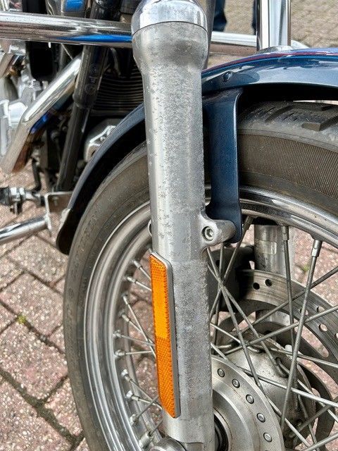 Nahaufnahme des Vorderrads eines Motorrads. Zu sehen ist eine Chromgabel mit einem orangefarbenen Reflektor und einem dunkelblauen Kotflügel.