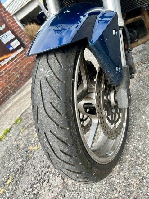 Nahaufnahme eines blauen Motorradvorderrads und Kotflügels auf einer Schotterfläche, schräg in der Nähe einer Backsteinmauer aufgenommen.