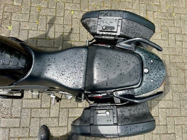 Draufsicht auf ein schwarzes Motorrad, das auf nassen Pflastersteinen geparkt ist; am unteren Rand sind die Schuhe einer Person zu sehen.