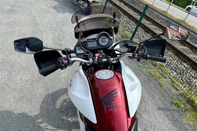 Eine Nahaufnahme des Lenkers, des Armaturenbretts und der Windschutzscheibe eines weiß-roten Honda-Motorrads, das auf Asphalt neben einem Zaun geparkt ist.