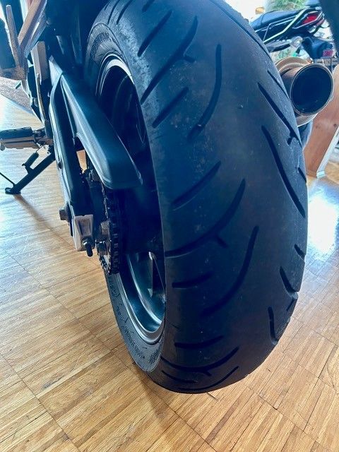Rückansicht eines Motorradreifens mit Felge auf einem Holzboden, Auspuff ist sichtbar.