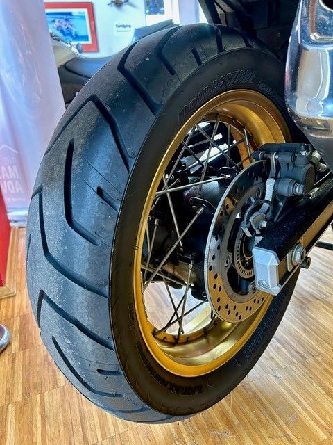 Nahaufnahme eines Motorradhinterrads mit goldener Felge, Scheibenbremse und dickem Reifen in einem Innenraum.