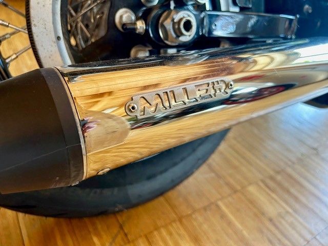 Verchromtes Motorradauspuffrohr mit Miller-Logo, Nahaufnahme eines Fahrradrads und Holzbodens