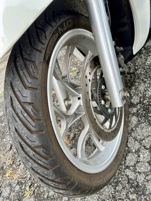 Nahaufnahme eines Motorrad-Vorderrads, Reifens und Bremsscheibe auf einer asphaltierten Fläche.