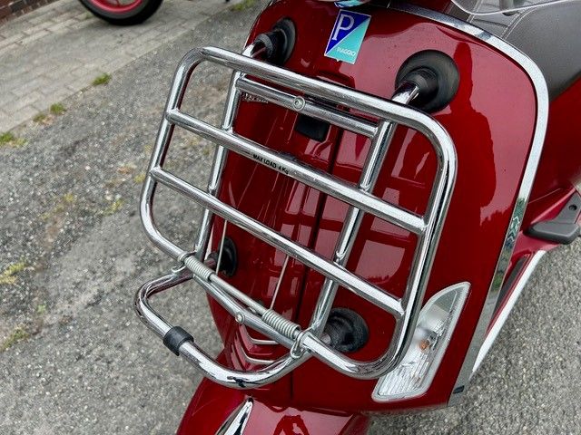 Ein roter Motorroller mit verchromtem Gepäckträger vorne steht am Straßenrand.