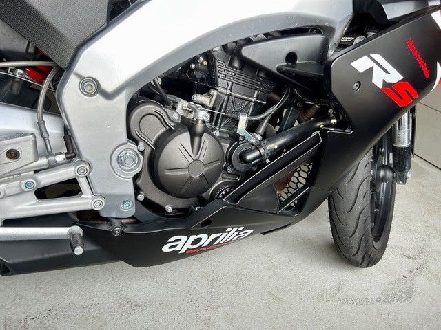 Nahaufnahme eines Aprilia-Motorradmotors und der Seitenverkleidung in Silber, Schwarz und Rot.