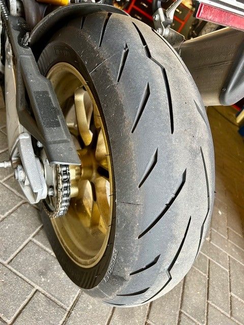 Nahaufnahme eines Motorradhinterrads mit goldener Felge und abgefahrenem Profil auf einem gefliesten Boden.