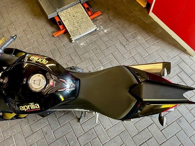 Draufsicht auf ein schwarzes Aprilia-Motorrad, das auf grauen Pflastersteinen in der Nähe einer roten Mauer geparkt ist.