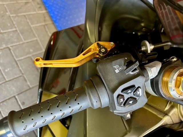 Nahaufnahme eines Motorradlenkers mit grauem Griff und goldenem Bremshebel an einem dunkelgrünen Rahmen.
