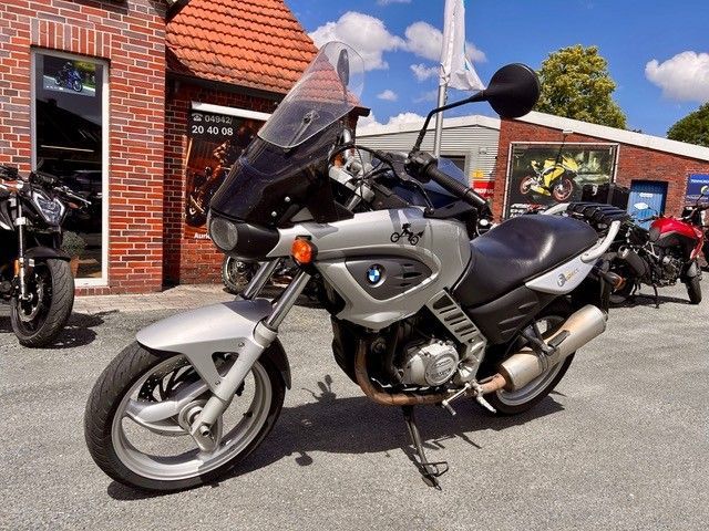 Ein silbernes BMW-Motorrad parkt an einem sonnigen Tag vor einem Backsteingebäude.