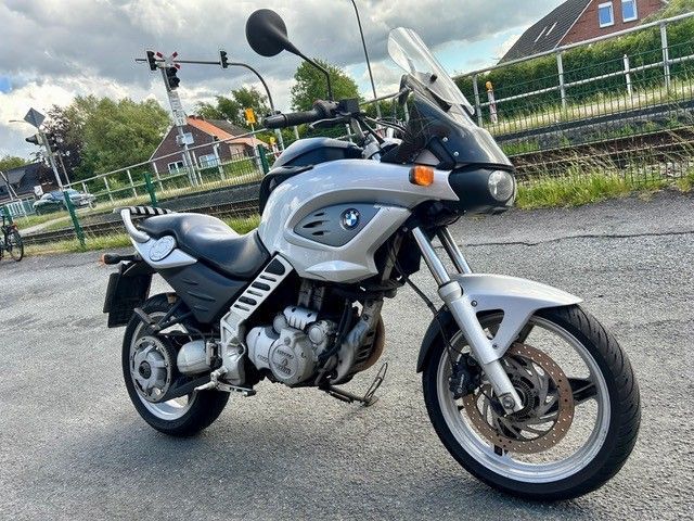 Ein silbernes BMW-Motorrad parkt im Freien neben einem Kanal, im Hintergrund sind Häuser zu sehen.