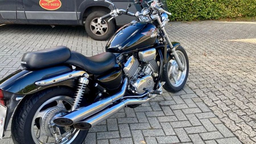 Schwarzes Honda-Motorrad mit Chromauspuff auf einer Ziegelfläche in der Nähe eines Lieferwagens geparkt.