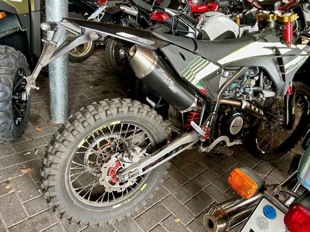 Dirtbike in einer Garage, Hinterrad und Auspuff sichtbar, Werkzeug und andere Fahrräder in der Nähe.
