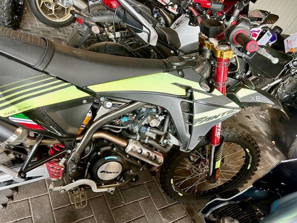Ein grün-schwarzes Dirtbike parkt auf einem Backsteinpflaster, im Hintergrund sind weitere Motorräder zu sehen.