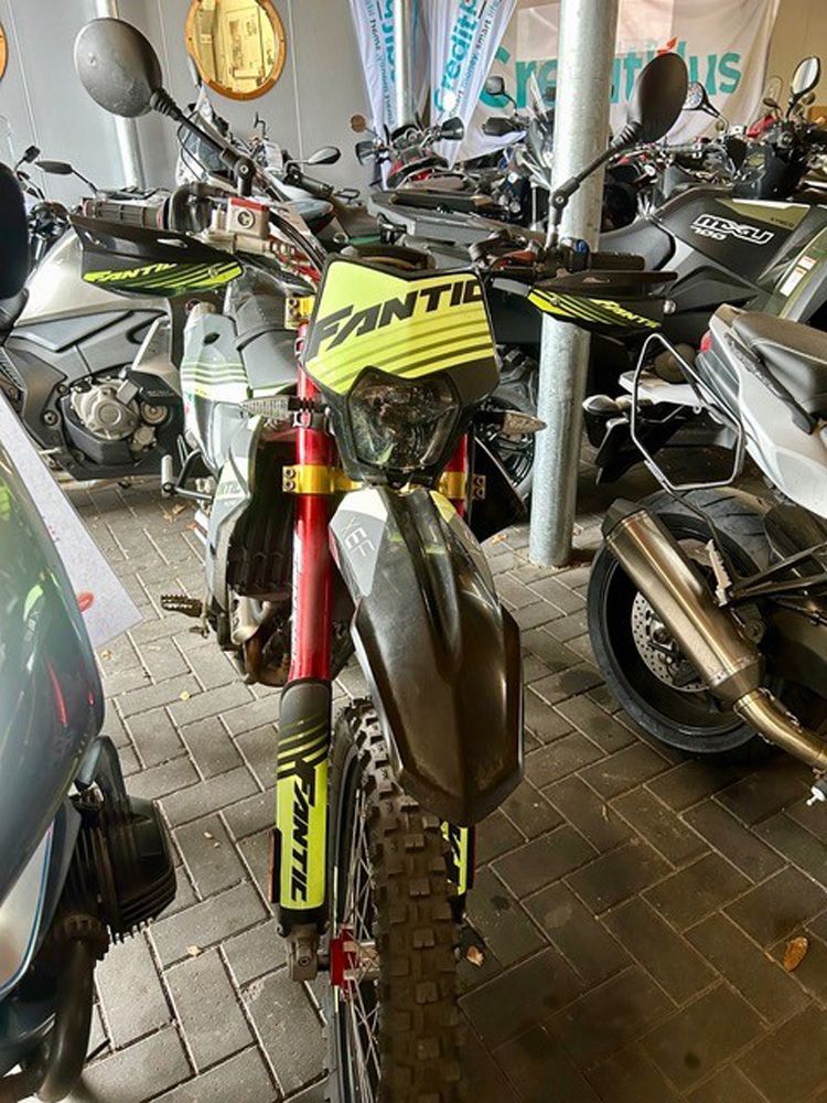Ein leuchtend gelbes Geländemotorrad steht in einem Innenraum zwischen anderen Motorrädern und Rollern.