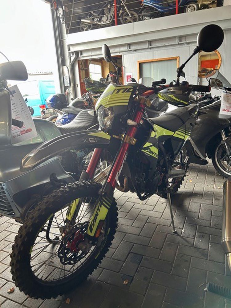 Ein Offroad-Dirtbike mit roter Gabel und Neonakzenten steht drinnen auf einem gefliesten Boden.
