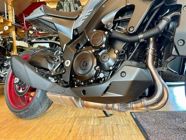 Nahaufnahme eines schwarzen Sportmotorrads mit roten Felgen auf einem Holzboden, die den Motor und den Auspuff zeigt.