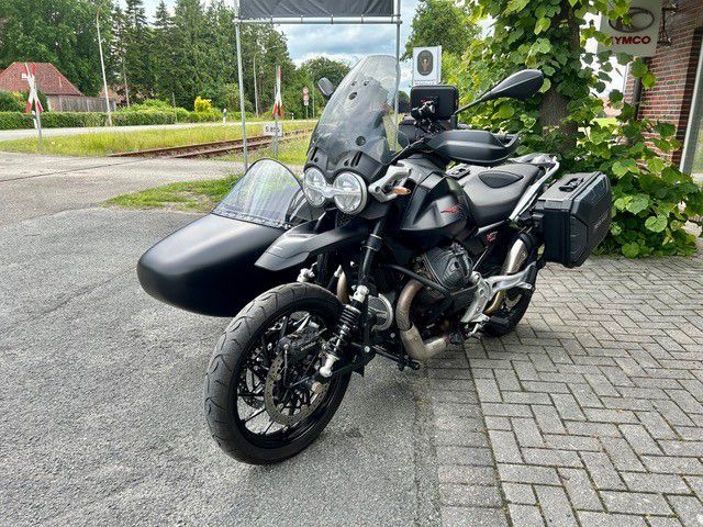 Schwarzes Tourenmotorrad mit Beiwagen, geparkt auf einer gepflasterten Auffahrt in der Nähe von Grünflächen und einem Gebäude.