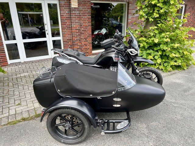 Schwarzes Motorrad mit Beiwagen parkt vor einem Backsteingebäude mit Fenstern und Grünflächen.