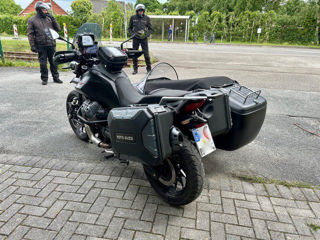 Ein dreirädriges Tourenmotorrad steht auf einem gepflasterten Parkplatz; zwei Personen stehen dahinter.