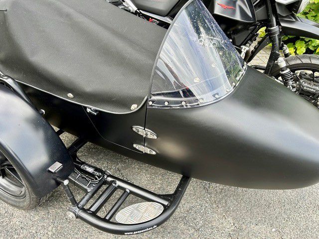 Schwarzer Motorradbeiwagen mit klarer Windschutzscheibe und schwarzer Abdeckung, geparkt auf dem Bürgersteig.