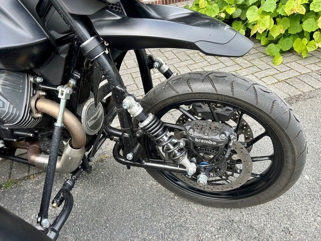 Schwarzes Motorrad parkt auf dem Bürgersteig neben grünen Sträuchern; Hinterrad und Auspuff sind zu sehen.