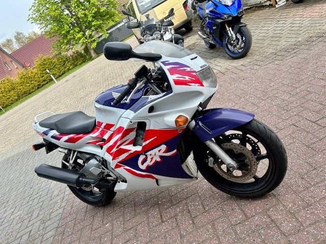 Ein blau-weißes Sportmotorrad mit roten Grafiken parkt auf einer gepflasterten Straße; im Hintergrund sind weitere Motorräder zu sehen.