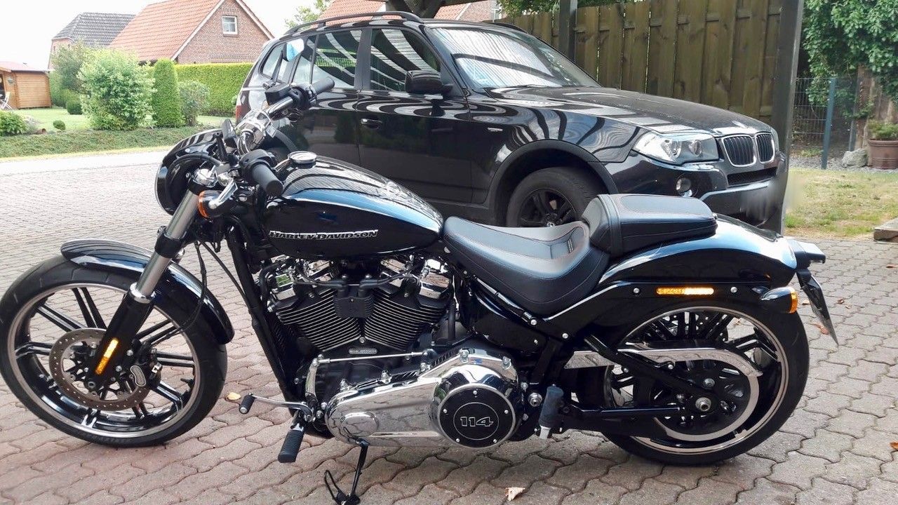 Ein schwarzes Cruiser-Motorrad parkt auf einer gepflasterten Auffahrt vor einem schwarzen Auto und einem Holzzaun.
