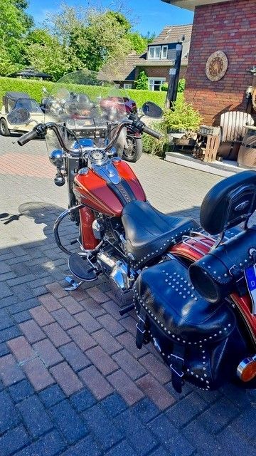 Ein rot-schwarzes Motorrad parkt auf einer gepflasterten Auffahrt neben einem Backsteinhaus; im Hintergrund ist ein Garten zu sehen.