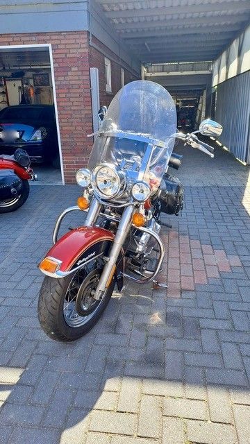 Ein rotes Cruiser-Motorrad mit hoher Windschutzscheibe parkt in einer gepflasterten Auffahrt unter einem Carport.