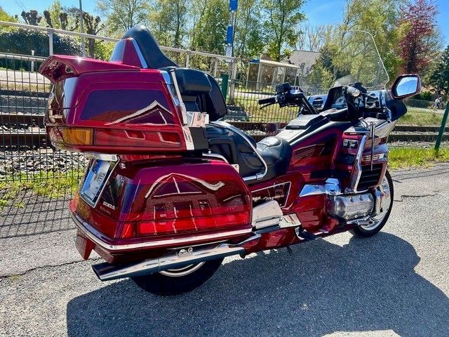 Ein rot-blaues Tourenmotorrad parkt an einem sonnigen Straßenrand in der Nähe eines Zauns und Bäumen.
