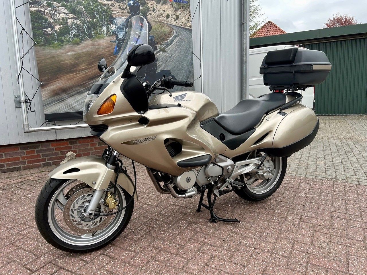 Beiges Tourenmotorrad mit Heckbox parkt auf einer gepflasterten Auffahrt neben einem Gebäude.