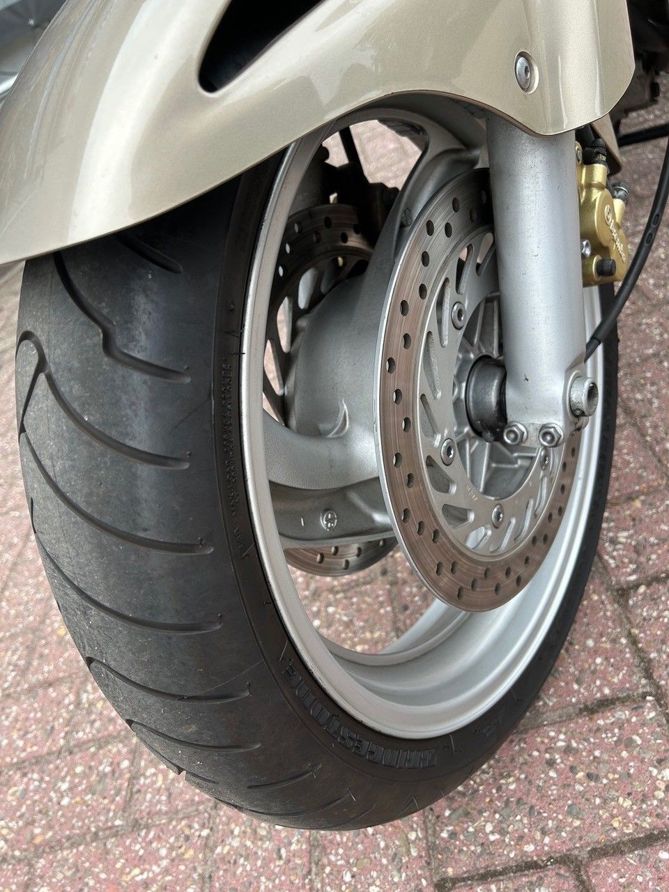 Nahaufnahme des Vorderrads und der Scheibenbremse eines Rollers auf einer gepflasterten Fläche.