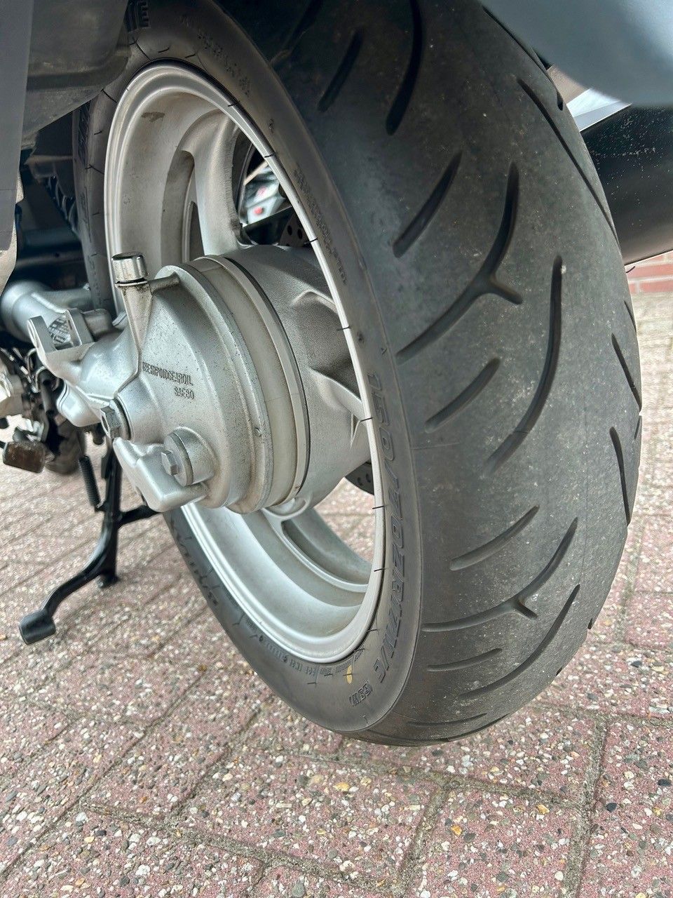 Nahaufnahme eines Motorradhinterrads mit Reifen auf asphaltierter Fläche, die die Bremsnabe und das Reifenprofil zeigt.