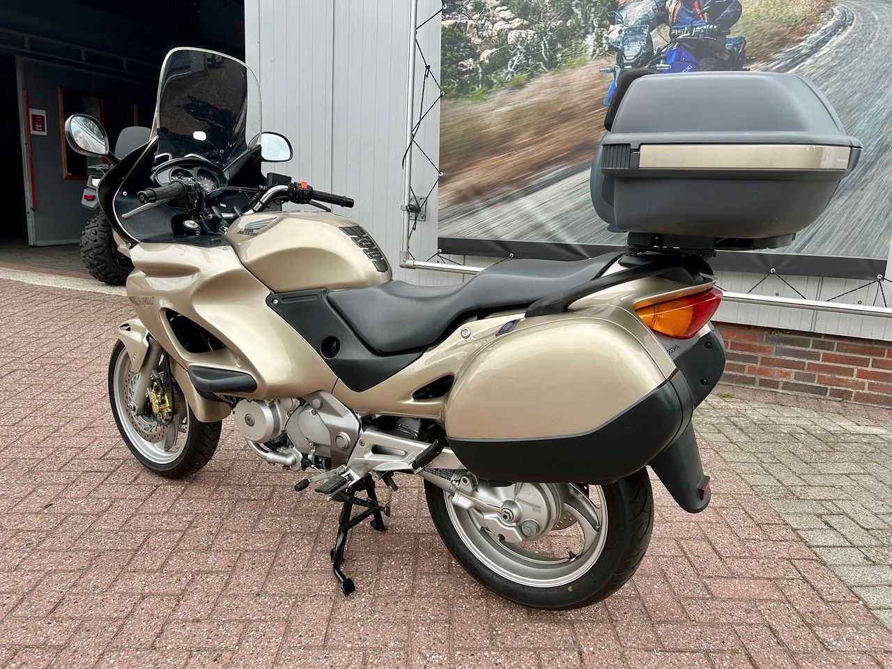 Ein beigefarbenes und schwarzes Tourenmotorrad parkt auf einem gepflasterten Gehweg vor einem Gebäude.