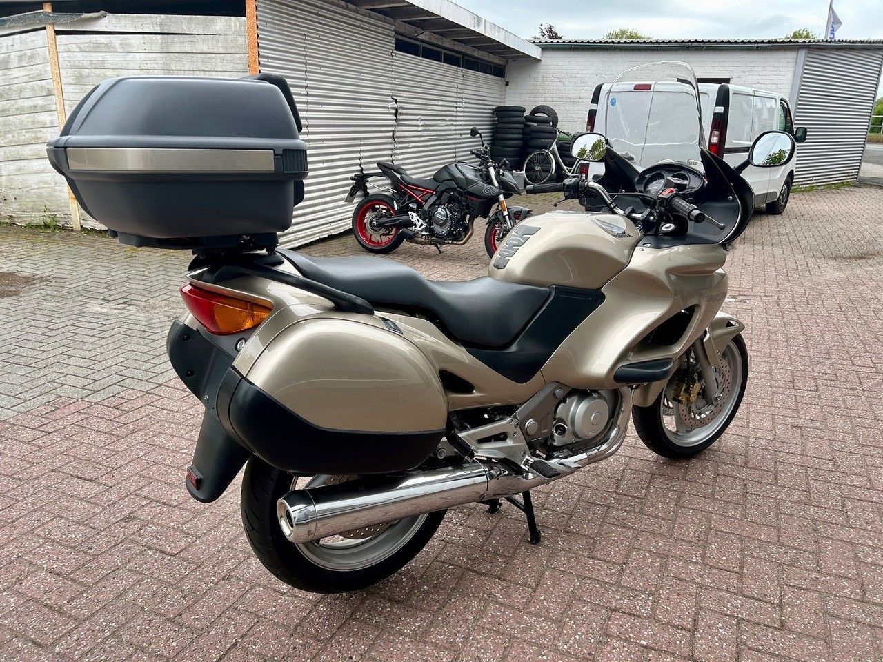 Beiges Tourenmotorrad mit Topcase parkt auf einer gepflasterten Auffahrt neben einem Wellblechgebäude.