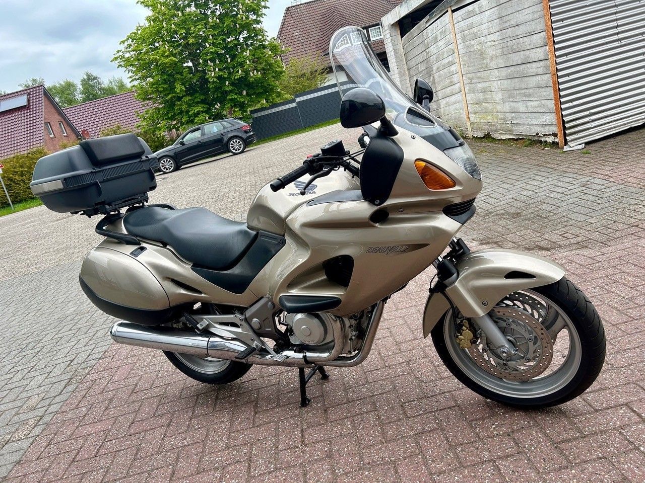 Ein goldenes Tourenmotorrad parkt auf einer gepflasterten Auffahrt neben einer Mauer und einer Garage.
