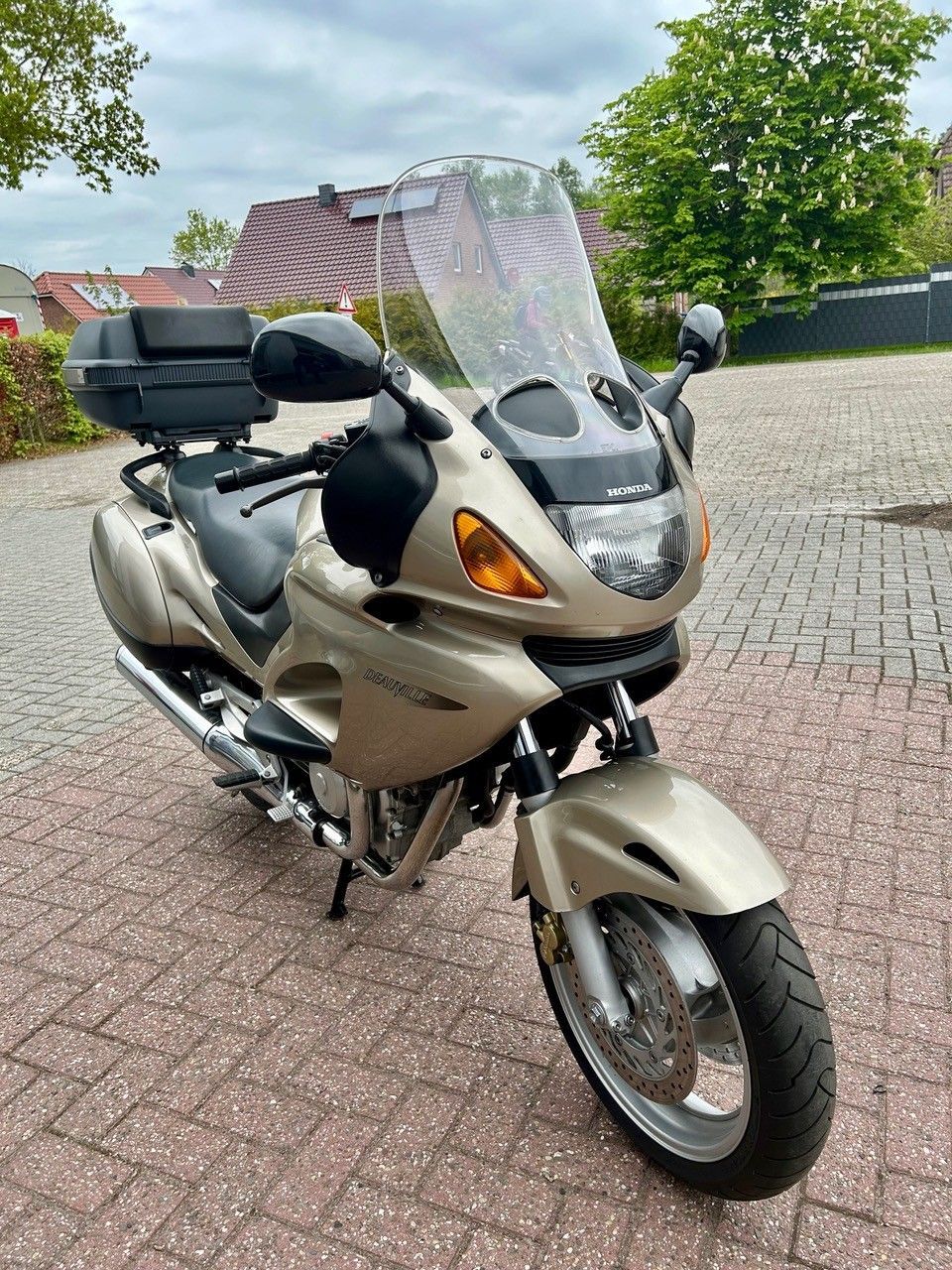 Silbernes Tourenmotorrad mit Windschutzscheibe und Topcase, geparkt auf einer gepflasterten Auffahrt.