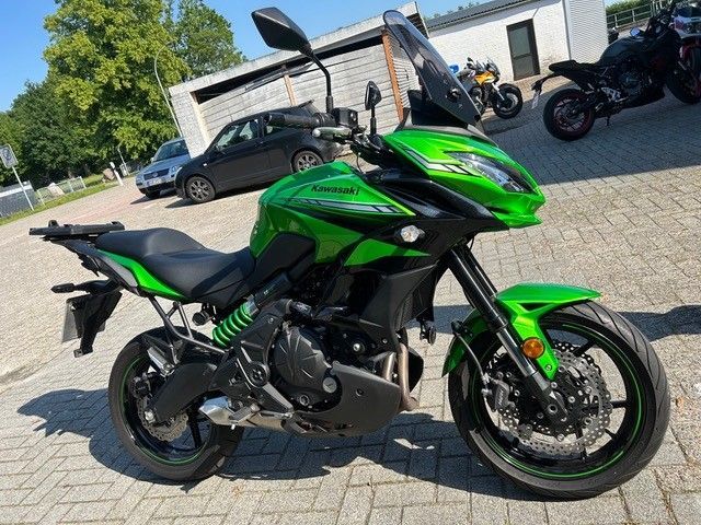 Ein grünes Sportmotorrad steht auf einem gepflasterten Parkplatz; im Hintergrund sind Gebäude und andere Motorräder zu sehen.