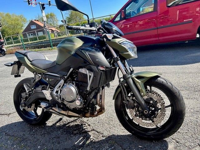 Ein mattschwarzes Sportmotorrad parkt in einer sonnigen Straße neben einem roten Lieferwagen und einem Haus.