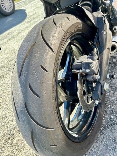 Nahaufnahme eines Motorradhinterreifens mit abgefahrenem Profil und schwarzer Felge auf einer sonnenbeschienenen Straße.