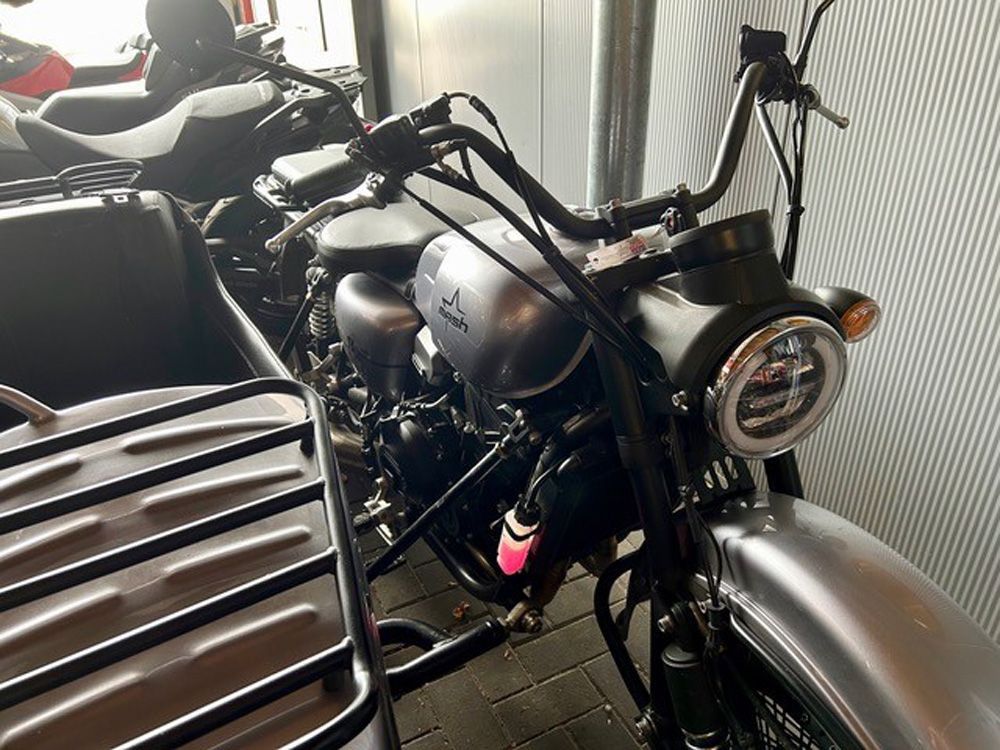 Schwarzes Motorrad parkt drinnen neben einer silbernen Bank und einer Wellblechwand, der Frontscheinwerfer ist auf die Kamera gerichtet.