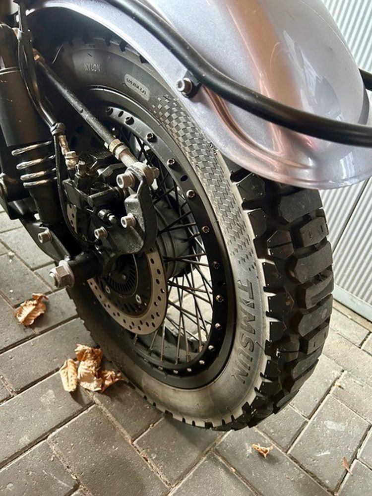 Nahaufnahme des Hinterrads und der Bremsscheibe eines Motorrads, das auf einem mit Laub bedeckten Backsteinpflasterweg parkt.