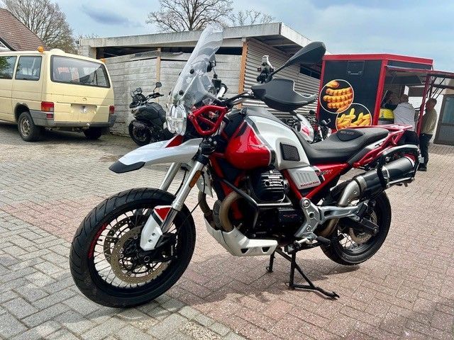 Ein rot-weißes Adventure-Motorrad parkt auf einer gepflasterten Straße neben einem Lieferwagen mit Anhänger.