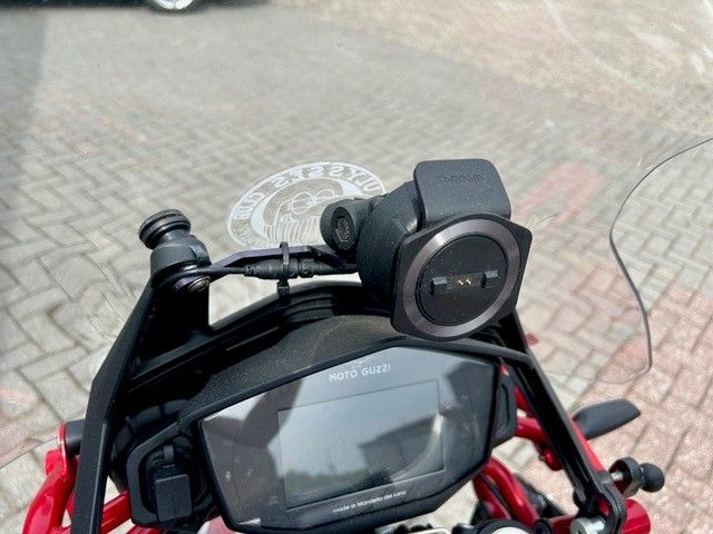 Motorradcockpit mit schwarzem Digitaldisplay und darüber montierter runder Kamera vor einem Backsteinpflaster-Hintergrund